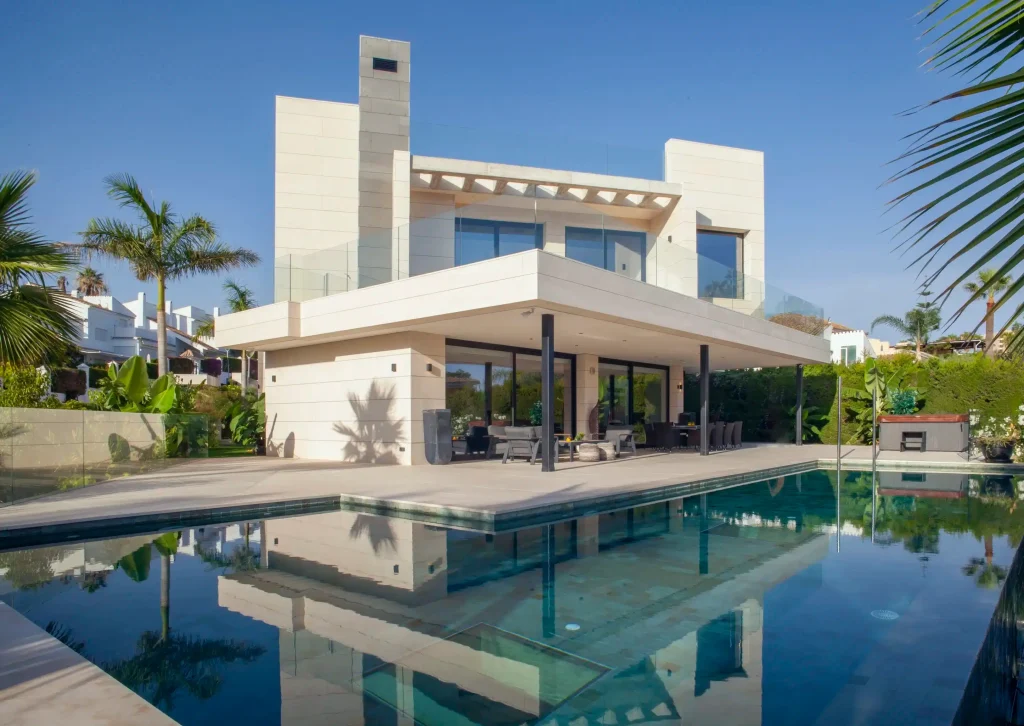 A modern luxury home with a swimming pool in the backyard, surrounded by lush greenery and illuminated by warm indoor lighting at sunset.